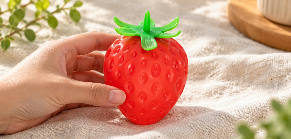 A hand holding a red strawberry-shaped squishy toy with a green leafy top on cream linen.